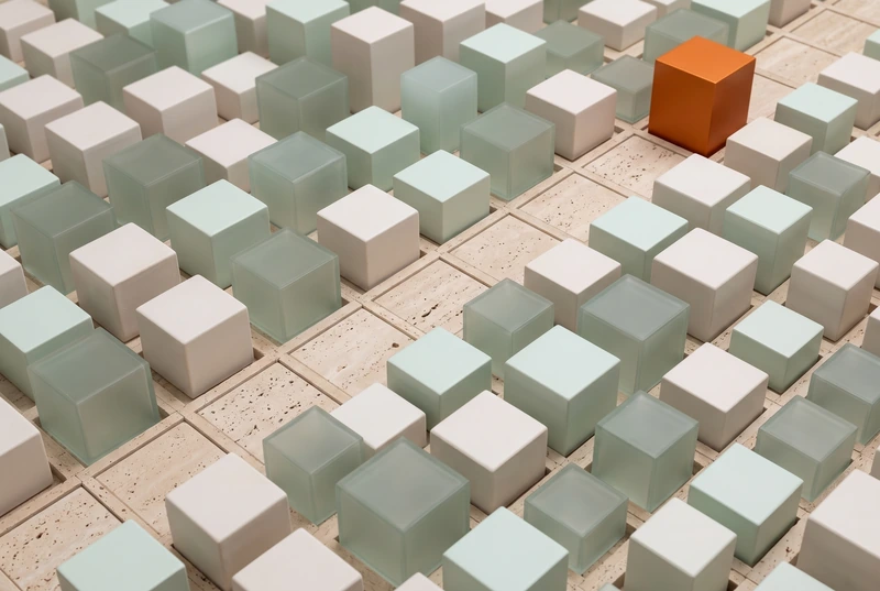 A travertine grid of glass and ceramic cubes with a clear path leading to one orange cube